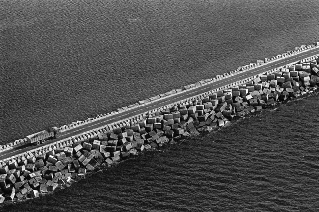 A road shored up with concrete blocks cuts diagonally across the frame, either side is a dark tone of sea. On this causeway a trck appears in the bottom left of the frame. The most noticeable part of the trcuk is the circles of the wheels, their roundness contrasting with the sharp edges of the blocks. The dark shadowy sides of the blocks meet bright sunlit sides, their strong shapes set against the soft texture of a dark sea.