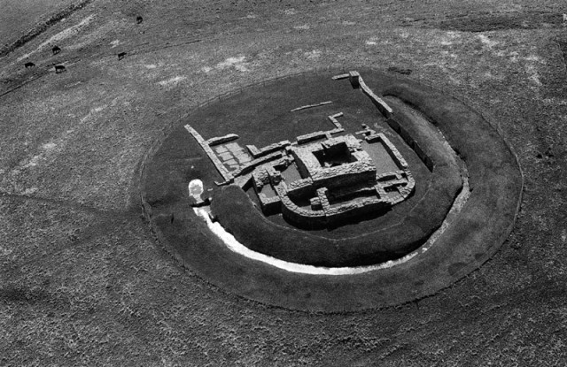 This black and white aerial photograph of the remaining ground floor of a former stronghold, shows the surrounding ditch encircling the curved walls and a dominant black square of a cellar and well central to the site. From above the archaeological site is a sketch in stone.