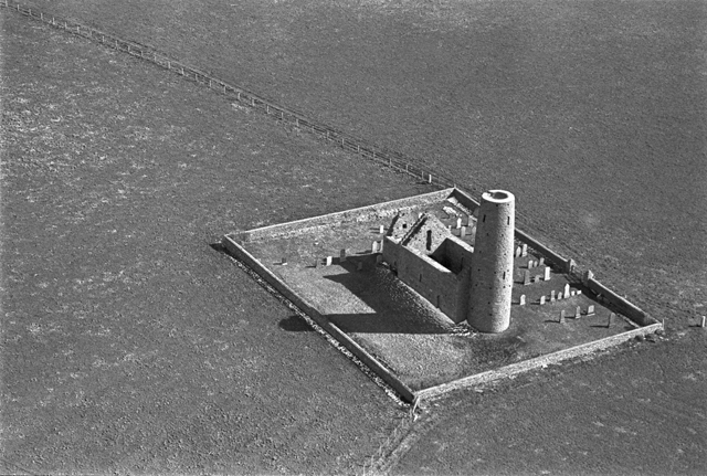 St Magnus Kirk on Egilsay photographed from the sky, a long shadow of the turret cuts across the kite shape creaed by the stone wall.